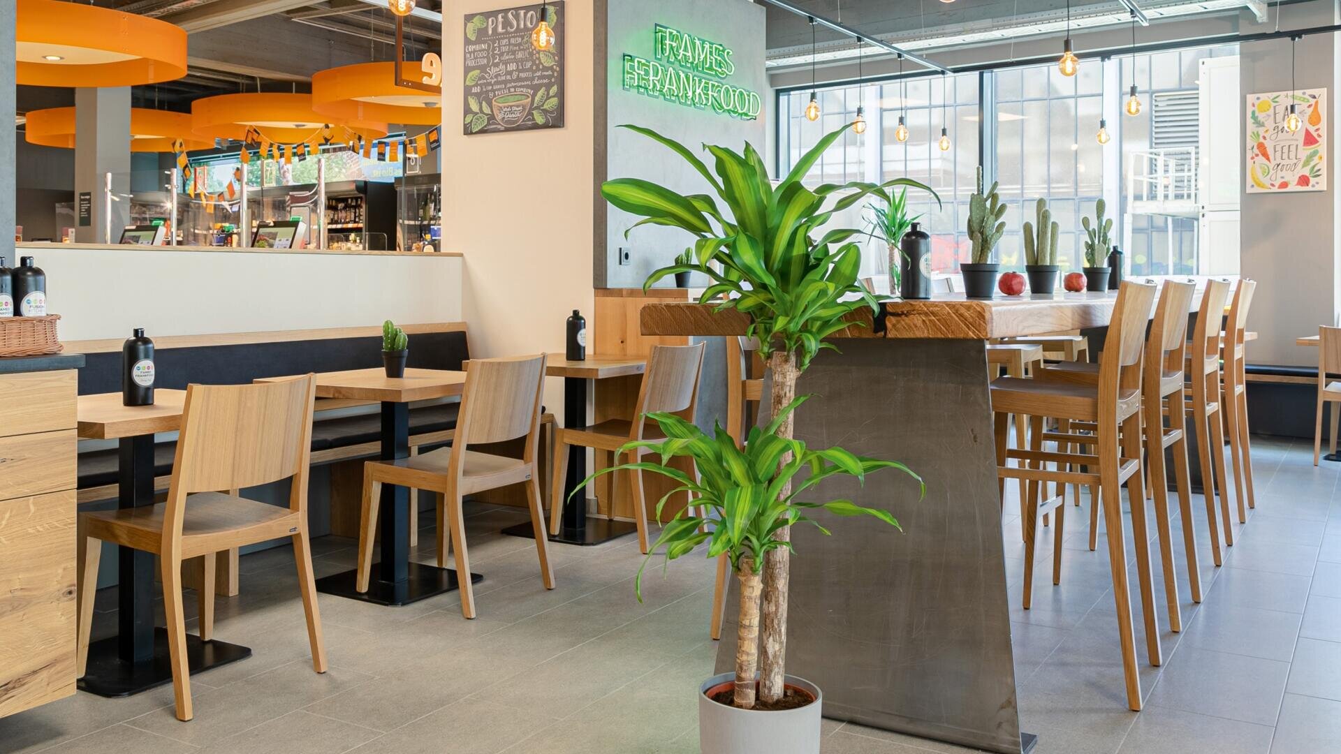 Un café moderne et lumineux avec des tables et des chaises en bois, un comptoir haut avec des tabourets de bar, des plantes vertes, de grandes fenêtres laissant entrer la lumière naturelle et une enseigne en néon indiquant FRAMES BEYOND FOOD (Cadres au-delà de la nourriture).