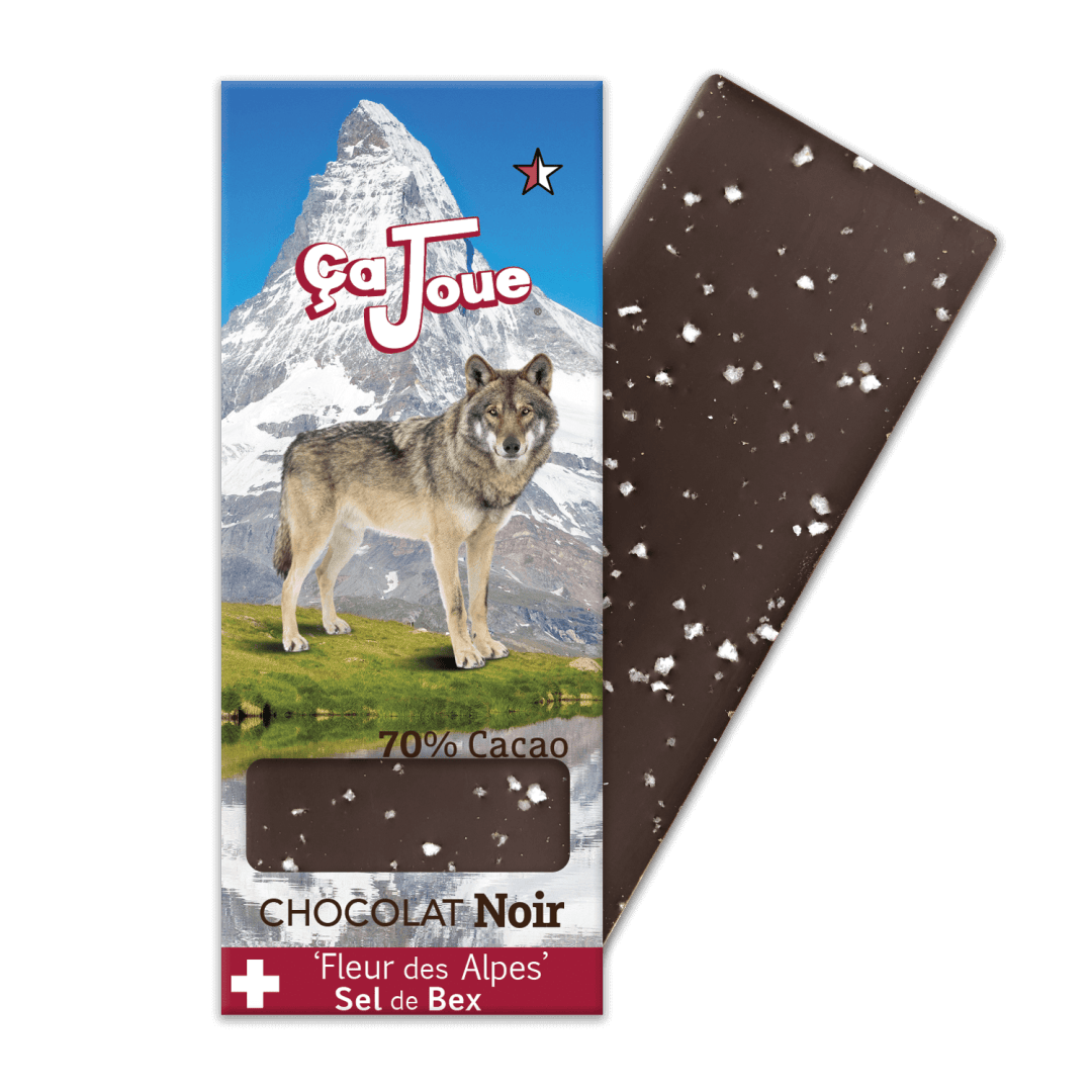 Une barre de chocolat avec des paillettes de sel visibles se trouve à côté de son emballage, qui présente un loup, un paysage de montagne et le texte suivant : Çà Joue, 70% Cacao, Chocolat Noir, et Fleur des Alpes Sel de Bex.