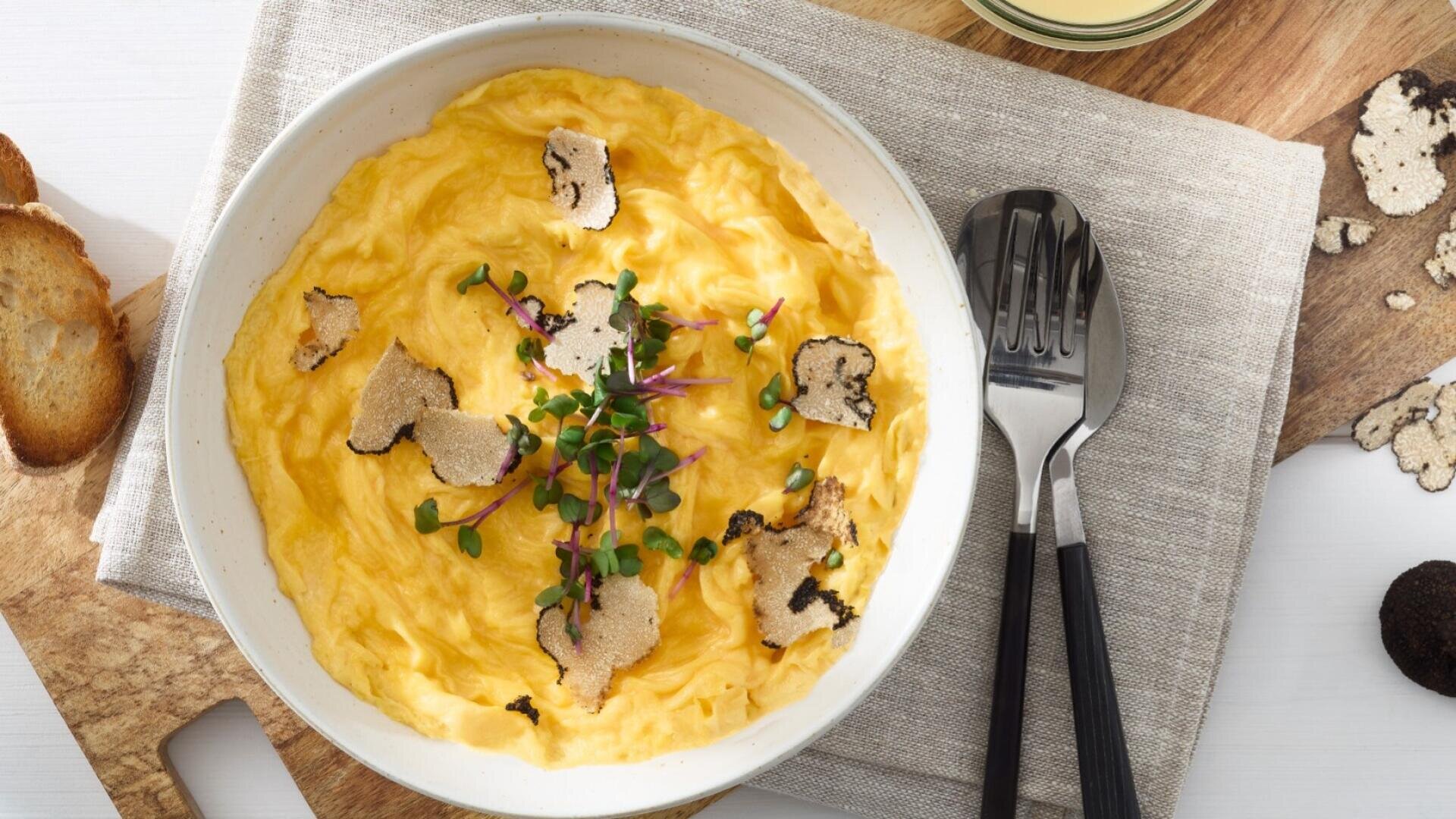 Eine Schale mit cremigem Eifix Schlemmer Rührei, garniert mit Kresse und schwarzen Trüffelscheiben, auf einer beigen Serviette, daneben Gabel und Löffel, daneben geröstetes Brot auf einem Holzbrett.