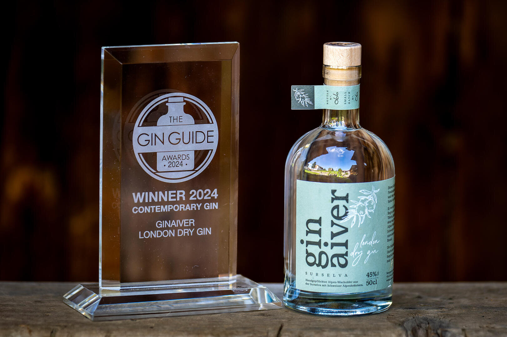 Eine Flasche Gin Aiver London Dry Gin steht neben einer Trophäe aus Klarglas mit der Aufschrift The Gin Guide Awards 2024 Winner: Zeitgenössischer Gin. Beide Gegenstände sind auf einer Holzfläche mit dunklem Hintergrund ausgestellt.