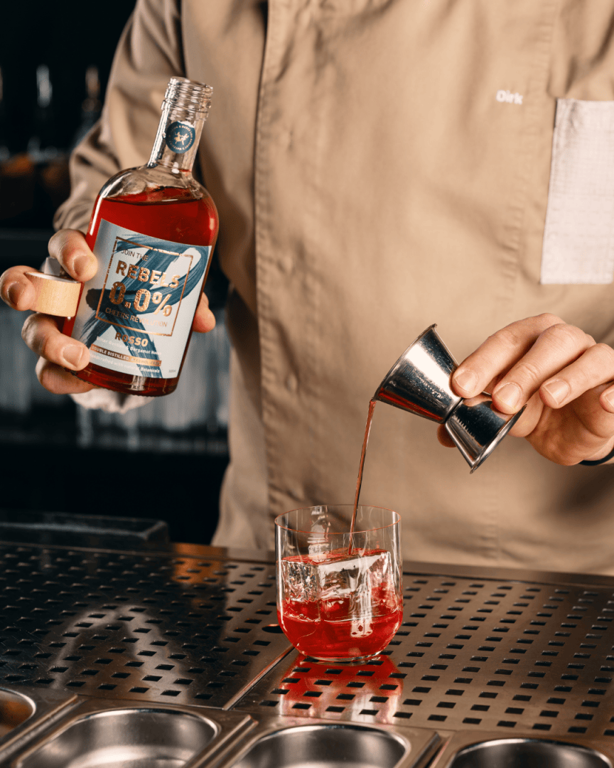 Ein Barkeeper gießt ein rotes Getränk aus einem Jigger in ein Glas mit Eis ein und hält eine Flasche mit der Aufschrift 0,0%. Der Barkeeper trägt ein beigefarbenes Hemd und steht hinter einem metallenen Bartresen.
