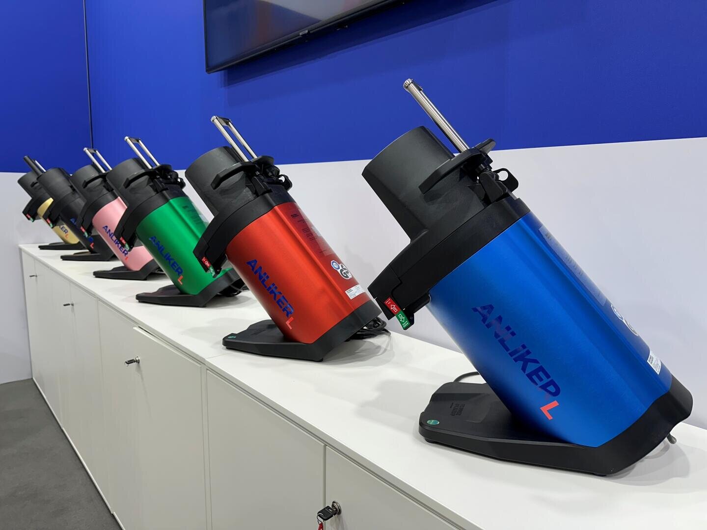 Plusieurs appareils cylindriques colorés portant l'inscription "AN-KHED" sont exposés en ligne sur une armoire blanche devant un mur bleu et blanc. Les appareils sont inclinés vers l'avant et apparaissent dans différentes couleurs : bleu, vert, rose, rouge et noir.