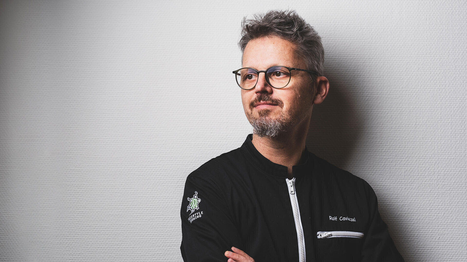 Ein Mann mit kurzen grauen Haaren, Brille und Bart steht vor einer hellen, strukturierten Wand. Er trägt eine schwarze Jacke mit einem Reißverschluss und einem Logo auf dem Ärmel und schaut nachdenklich zur Seite.