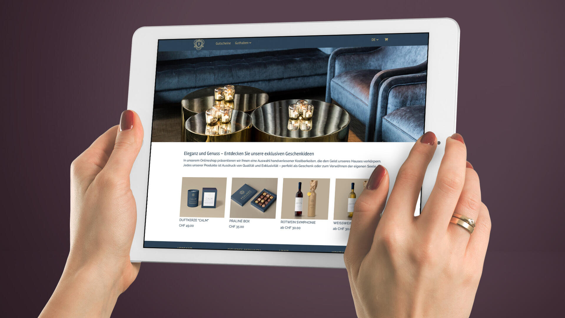Eine Person hält ein weißes Tablet, auf dem eine Luxus-Shopping-Website mit Bildern von goldfarbenen Schalen mit Snacks und Weinflaschen vor einem dunkelvioletten Hintergrund angezeigt wird.