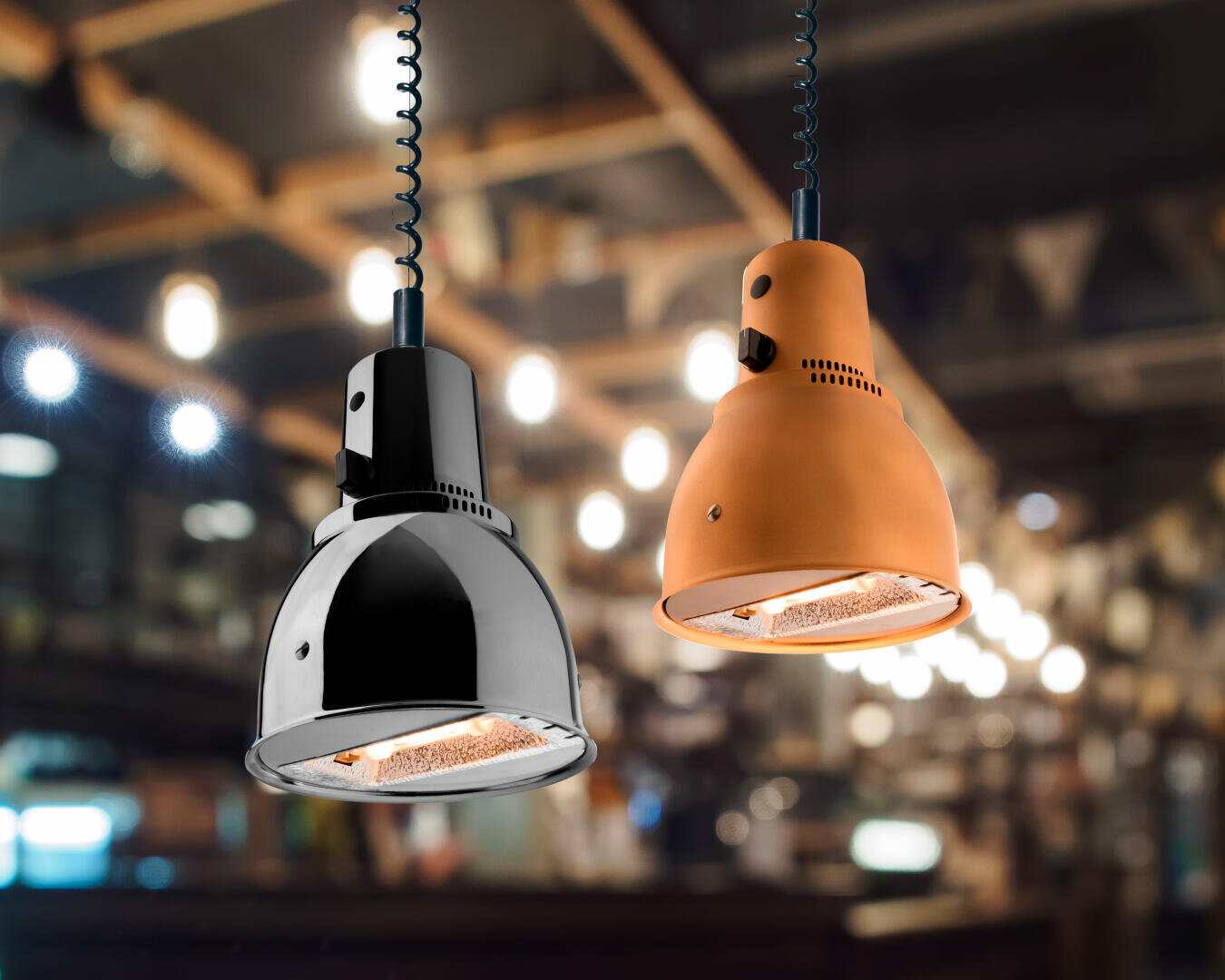 Zwei hängende Wärmelampen, eine metallisch-silberne und eine matt-orangefarbene, leuchten im Vordergrund eines warm beleuchteten, verschwommenen Restaurantinneren mit freiliegenden Balken und Lichterketten.