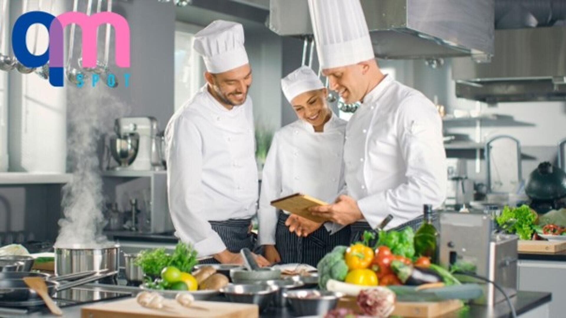Trois chefs en uniformes blancs et toques se tiennent dans une cuisine moderne, souriants et regardant une tablette numérique. Des légumes frais et des ingrédients sont posés sur le comptoir, tandis que la vapeur s'échappe d'une marmite. On distingue le logo QM Spot coloré.