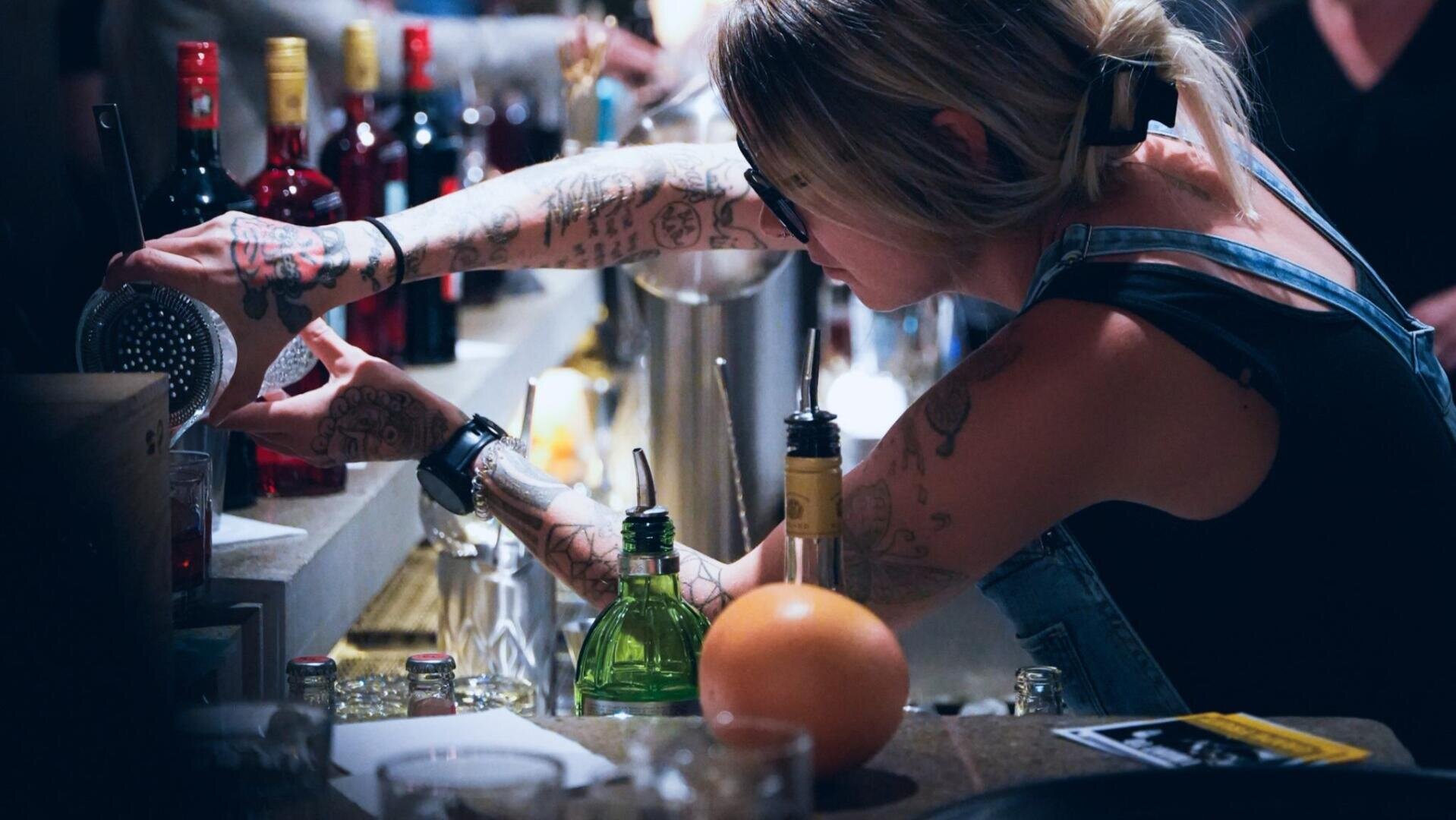 Derrière un bar, entourée de bouteilles, de shakers et d'un pamplemousse, une barmaid tatouée avec des lunettes et une queue de cheval sert un verre et se concentre sur sa tâche.