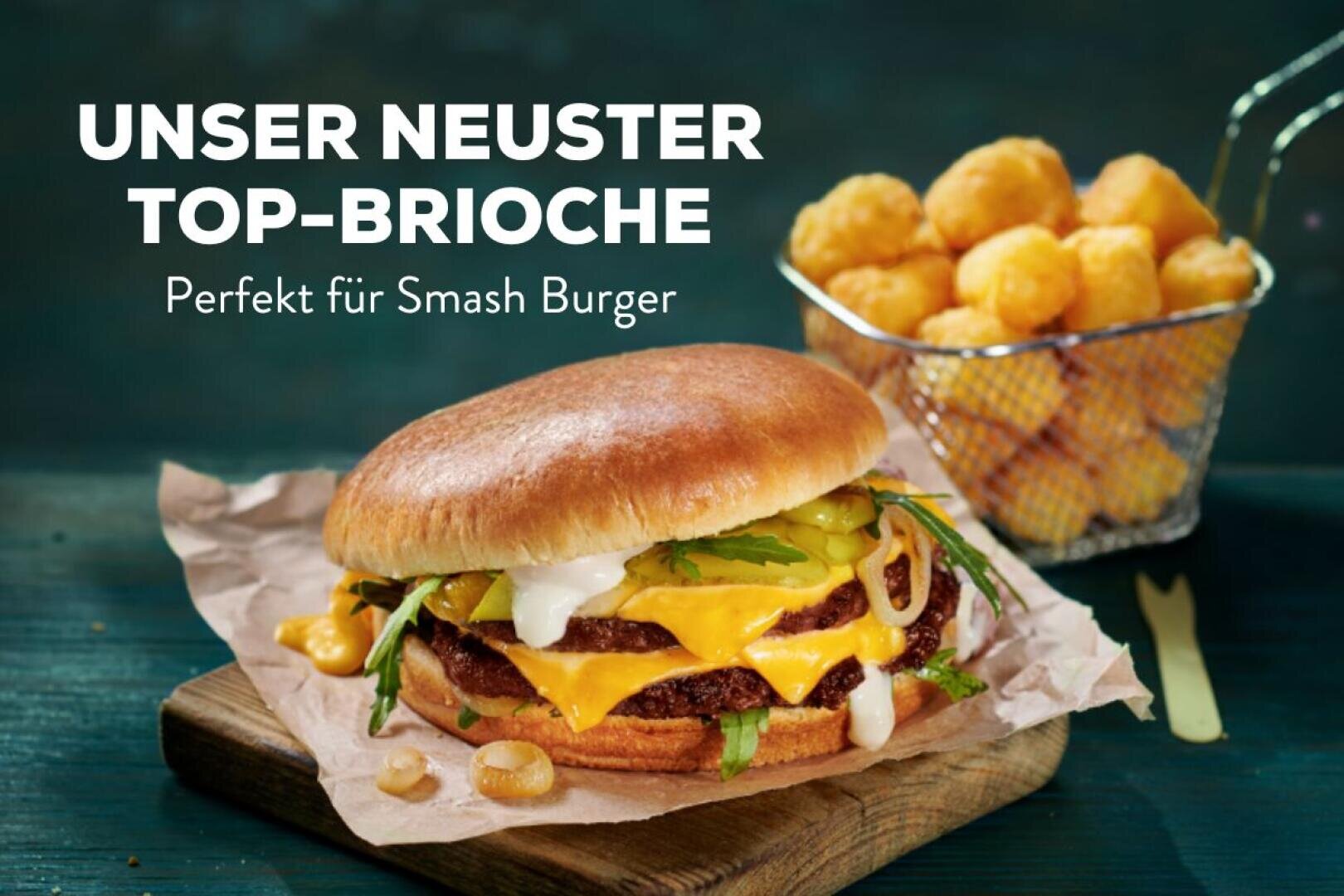 Ein Cheeseburger mit geschmolzenem Käse, Gurken, Zwiebeln und Sauce in einem Briochebrötchen liegt auf Backpapier. Im Hintergrund ist ein kleiner Korb mit goldenen Kartoffelecken zu sehen. Der Text lautet: „Unser neuster Top-Brioche. Perfekt für Smash Burger.“.
