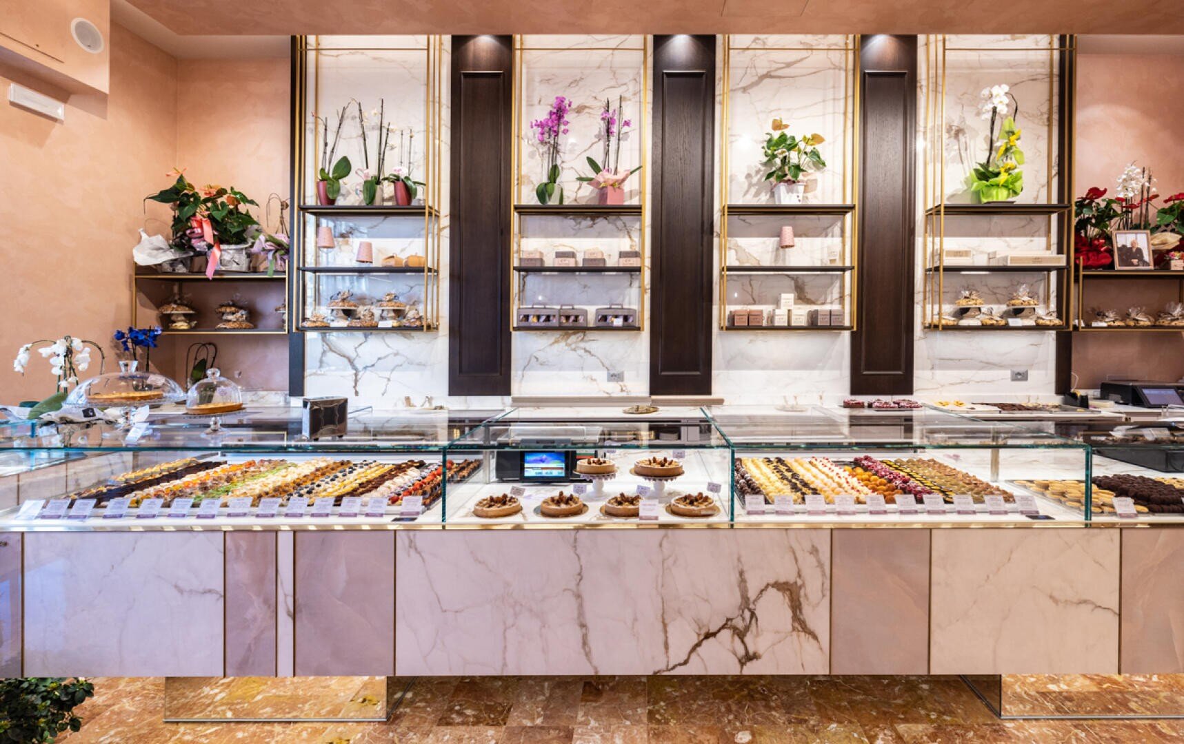 Eine stilvolle Konditorei mit einer Marmortheke, auf der verschiedene Backwaren und Pralinen angeboten werden. Hinter der Theke befinden sich elegante Regale mit Pflanzen, Blumen und Dekorationsartikeln, die eine schicke und einladende Atmosphäre schaffen.