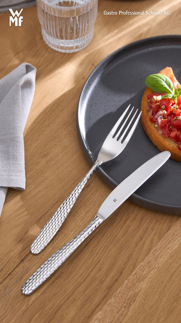 Une fourchette et des couteaux en argent aux manches texturés sont posés sur une table en bois à côté d'une assiette grise de bruschetta, d'un verre d'eau et d'une serviette beige. La lumière du soleil projette des ombres douces sur la scène.