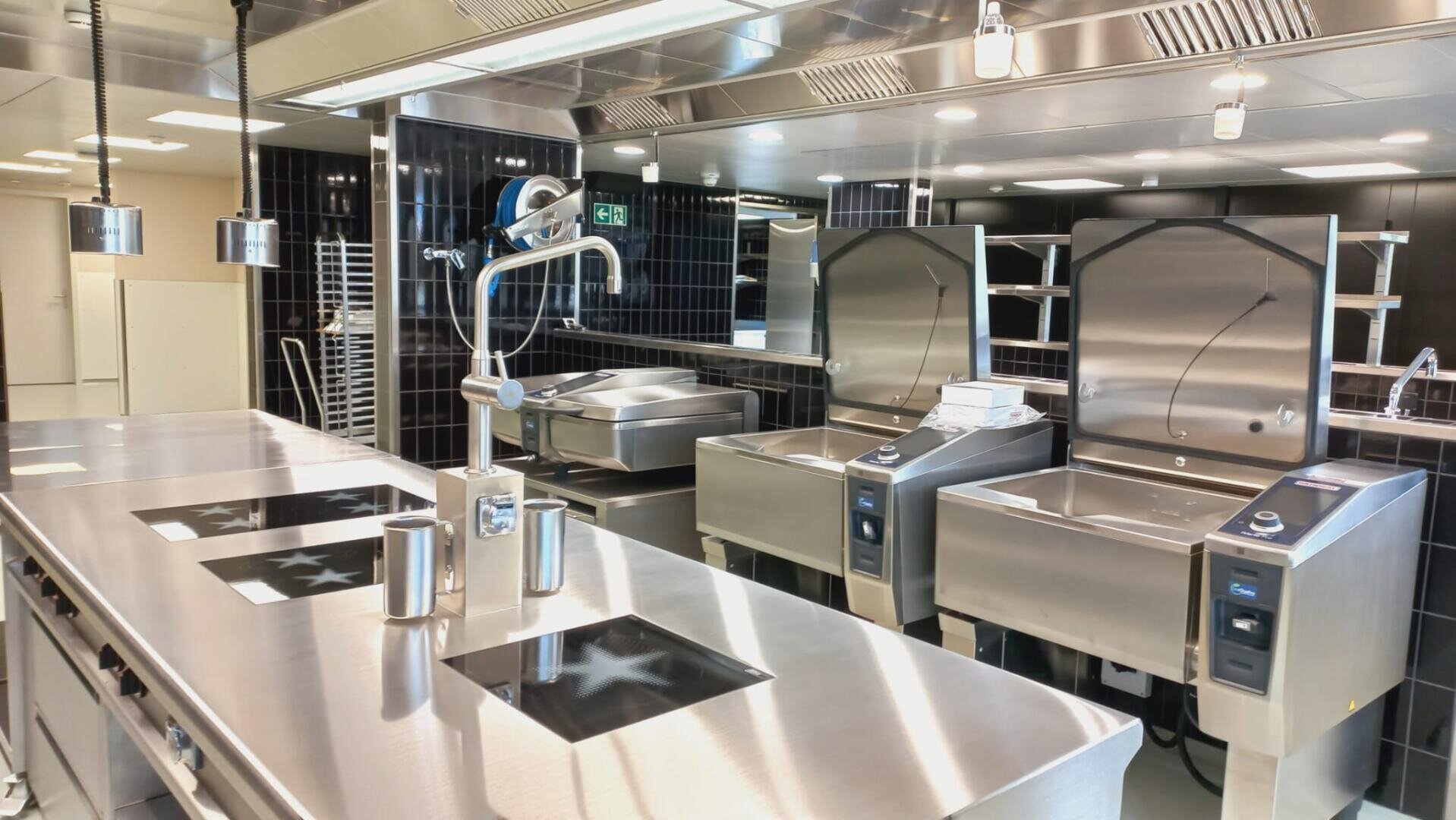 Eine moderne, professionelle Küche mit Edelstahloberflächen, Induktionskochfeldern, Backöfen, großen Kochgeräten und schwarzen Kachelwänden. Der Raum ist sauber, gut beleuchtet und für eine effiziente Essenszubereitung organisiert.