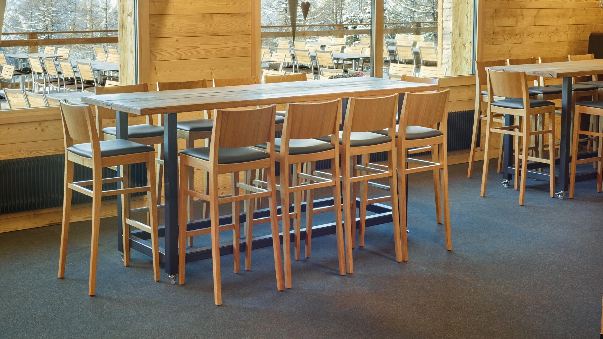Ein langer High-Top-Holztisch mit acht passenden Barhockern aus Holz in einem hellen, modernen Raum mit großen Fenstern und Sitzgelegenheiten im Freien im Hintergrund.