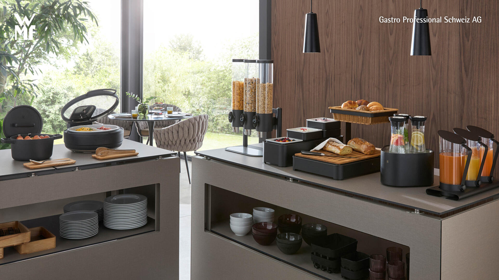 Un buffet de petit-déjeuner moderne avec des céréales, des pâtisseries, des fruits et des distributeurs de jus de fruits sur des comptoirs sobres. Les assiettes, les bols et les couverts sont clairement disposés. De grandes fenêtres donnent une vue sur la verdure.