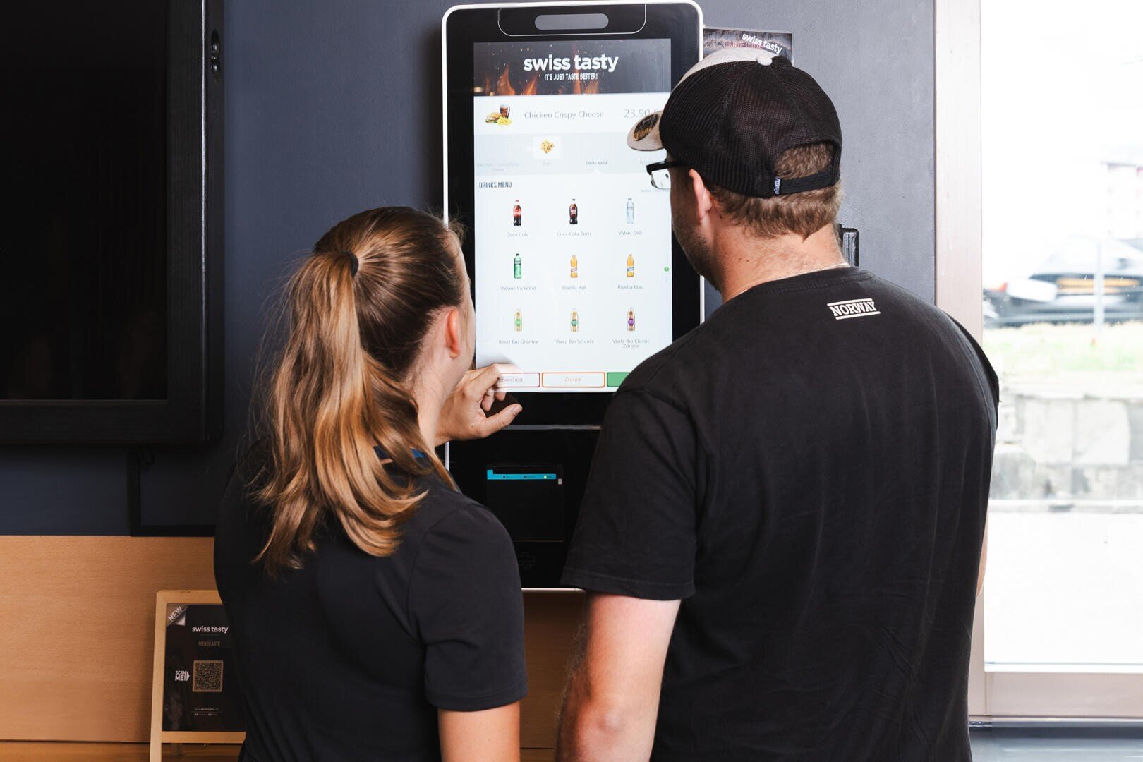 Zwei Personen stehen vor einem Touchscreen-Bestellkiosk in einem Restaurant und sehen sich gemeinsam die Menüoptionen auf dem Bildschirm an. Der Kiosk zeigt die Auswahl an Speisen und Getränken unter dem Restaurantnamen Swiss Tasty an.