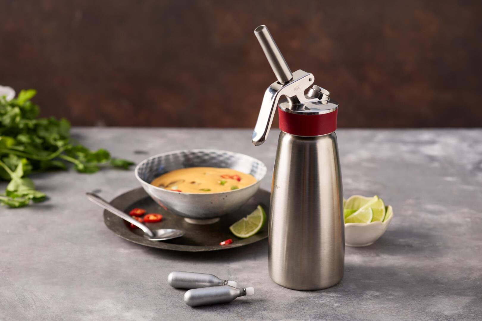 Ein Sahnespender aus Edelstahl und zwei Gaskartuschen stehen neben einer Schüssel Suppe, garniert mit roten Chilis und Kräutern, im Hintergrund auf einer grauen Fläche liegen Limettenspalten in einer kleinen Schale.