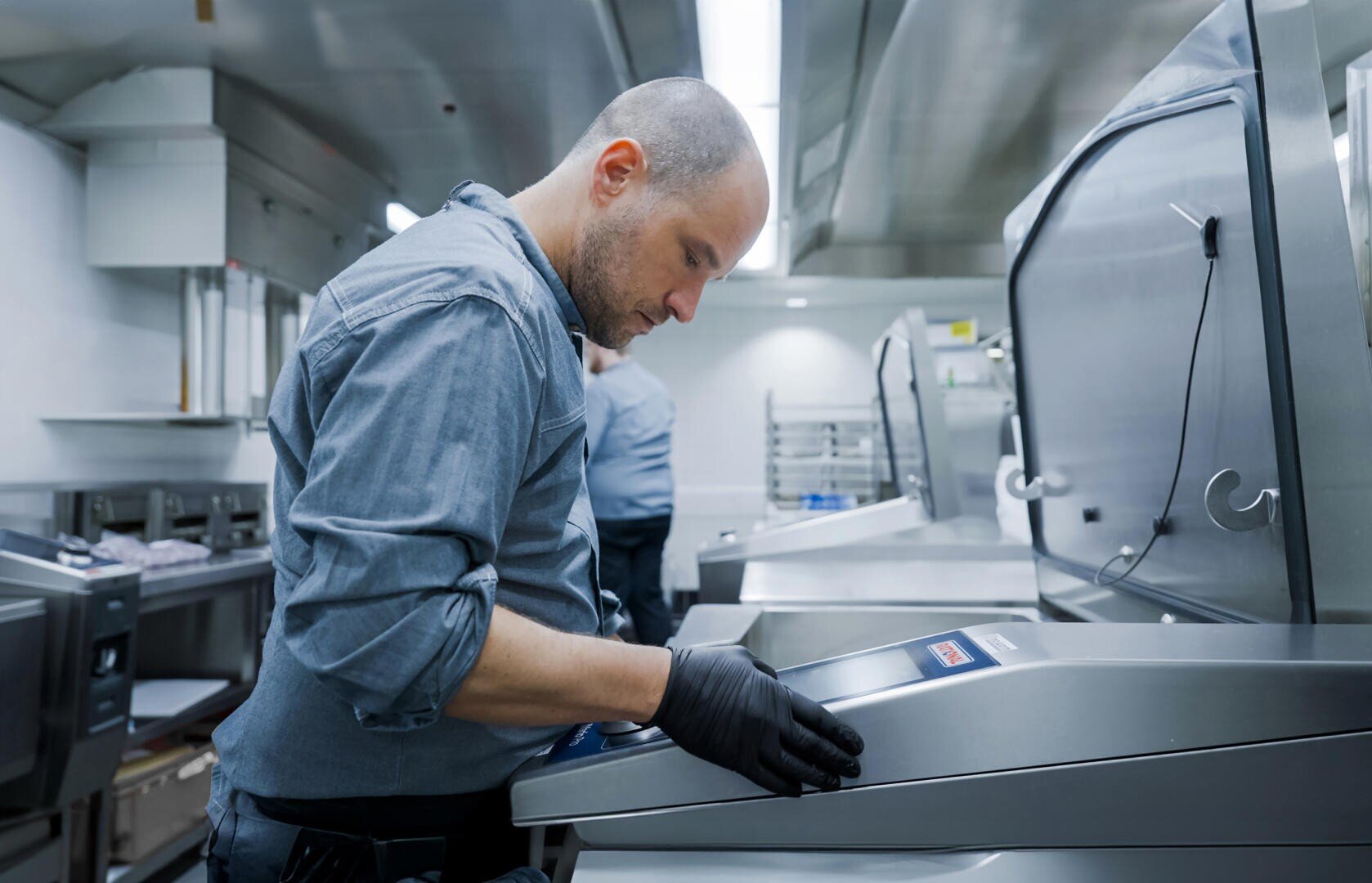 Ein Mann mit einem blauen Hemd und schwarzen Handschuhen bedient in einer Großküche eine große Maschine aus rostfreiem Stahl. Die Umgebung ist sauber, mit Industriegeräten und hellem Licht.