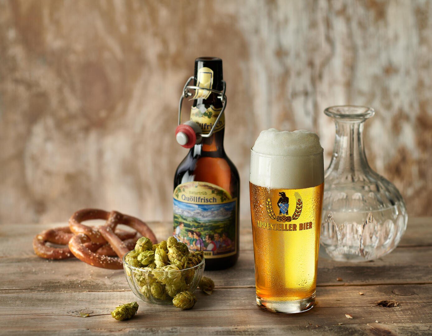 Ein Glas goldenes Bier mit Schaum, eine braune Flasche, eine klare Karaffe, eine Schale mit grünem Hopfen und zwei Brezeln stehen auf einem rustikalen Holztisch vor einem strukturierten Hintergrund.