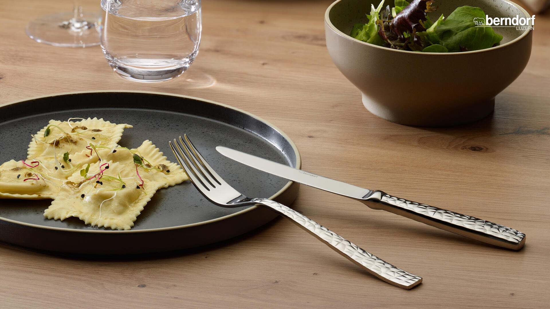 Auf einem Holztisch steht ein dunkler Teller mit vier Ravioli, die mit Sprossen belegt sind, sowie Gabel und Messer. Im Hintergrund sind auch ein Glas Wasser und eine Schüssel mit Salat zu sehen.