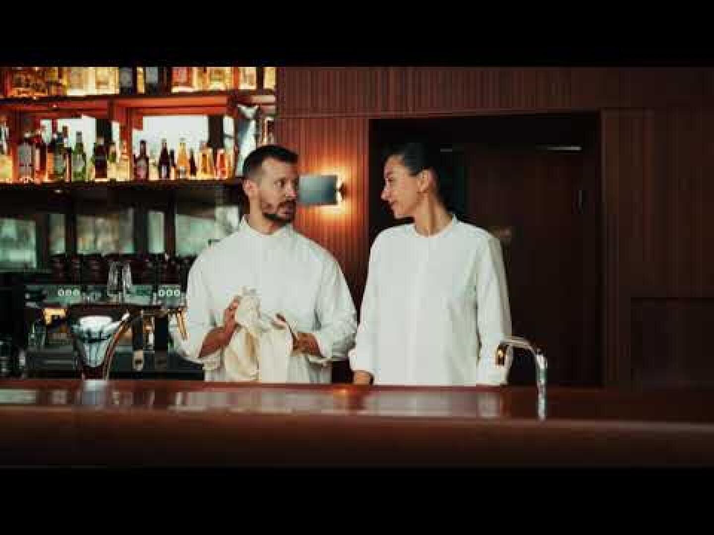 Zwei Barkeeper in weißen Uniformen stehen hinter einer Bar, lächeln und schauen sich an. Im Hintergrund sind Regale mit Flaschen zu sehen. Ein Barkeeper trocknet ein Glas mit einem Handtuch ab.