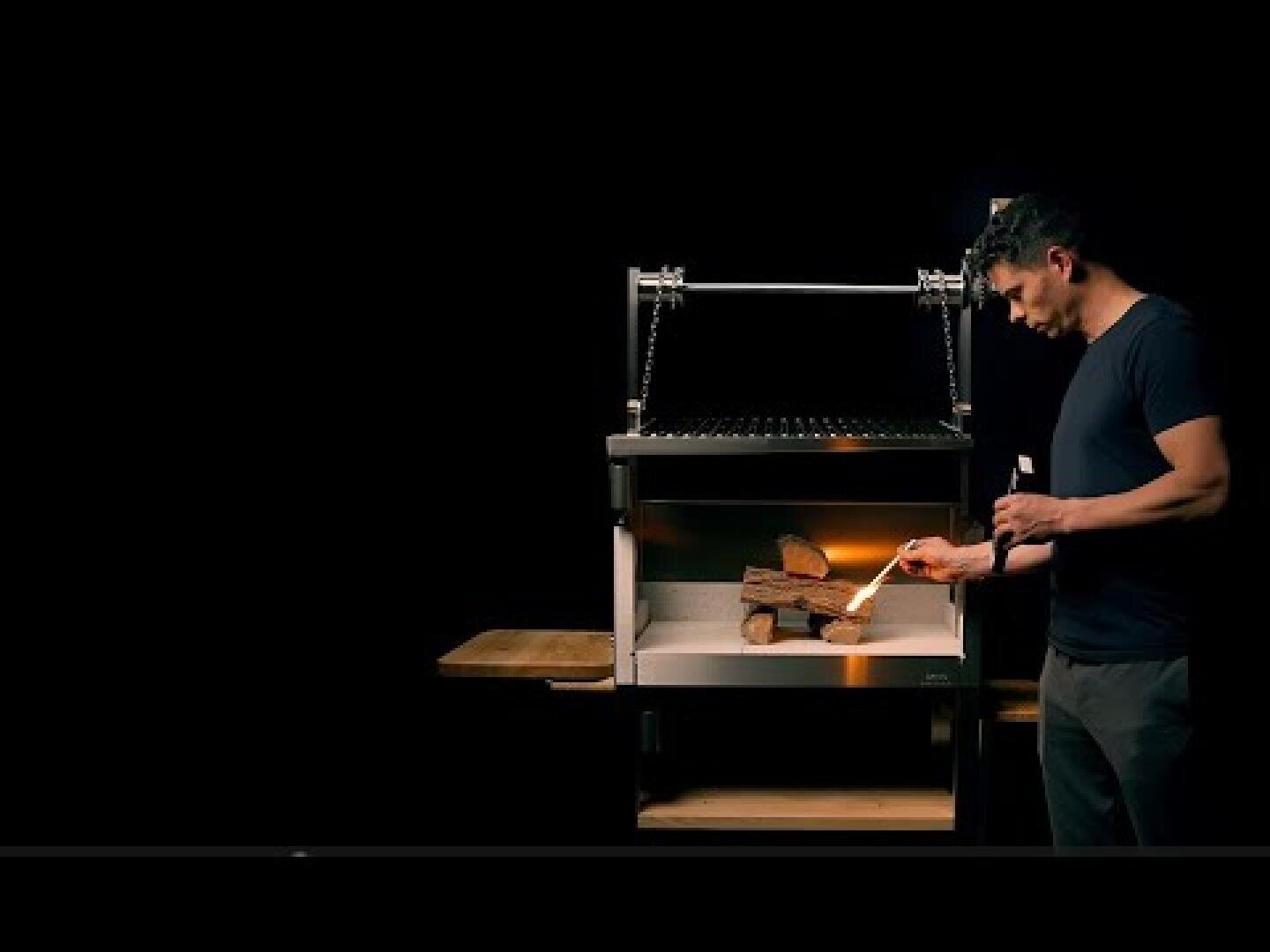 Ein Mann steht neben einem modernen Grill und entzündet das Holz darin mit einem Streichholz. In der anderen Hand hält er ein Getränk. Der Hintergrund ist dunkel, so dass die Szene und der Grill im Mittelpunkt des Bildes stehen.