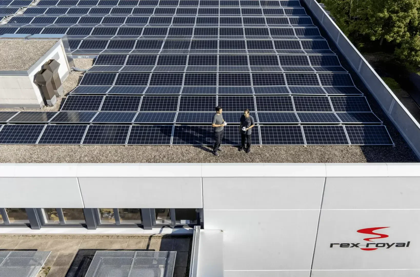 Luftaufnahme von zwei Personen, die Solarmodule auf einem großen flachen Dach eines weißen Gebäudes inspizieren, wobei das rex-royal-Logo und Bäume im Hintergrund zu sehen sind.