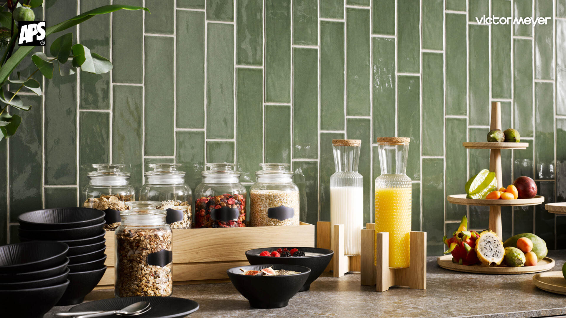 Un comptoir de cuisine avec des bols de céréales, des coupes de fruits, des petits bols noirs, un pichet en verre avec du lait, un pichet de jus d'orange et un présentoir à fruits devant un plan de travail carrelé vert.