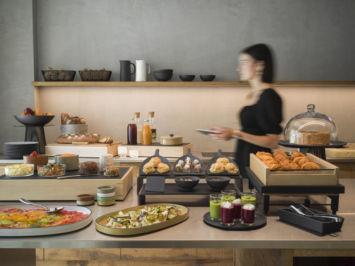 Ein Buffettisch mit Gebäck, Brot, Obst, Salaten, Säften und Gewürzen. Eine unscharfe Frau steht davor, hält einen Teller und bedient sich. Im Hintergrund sind Regale mit Geschirr und Gläsern zu sehen.