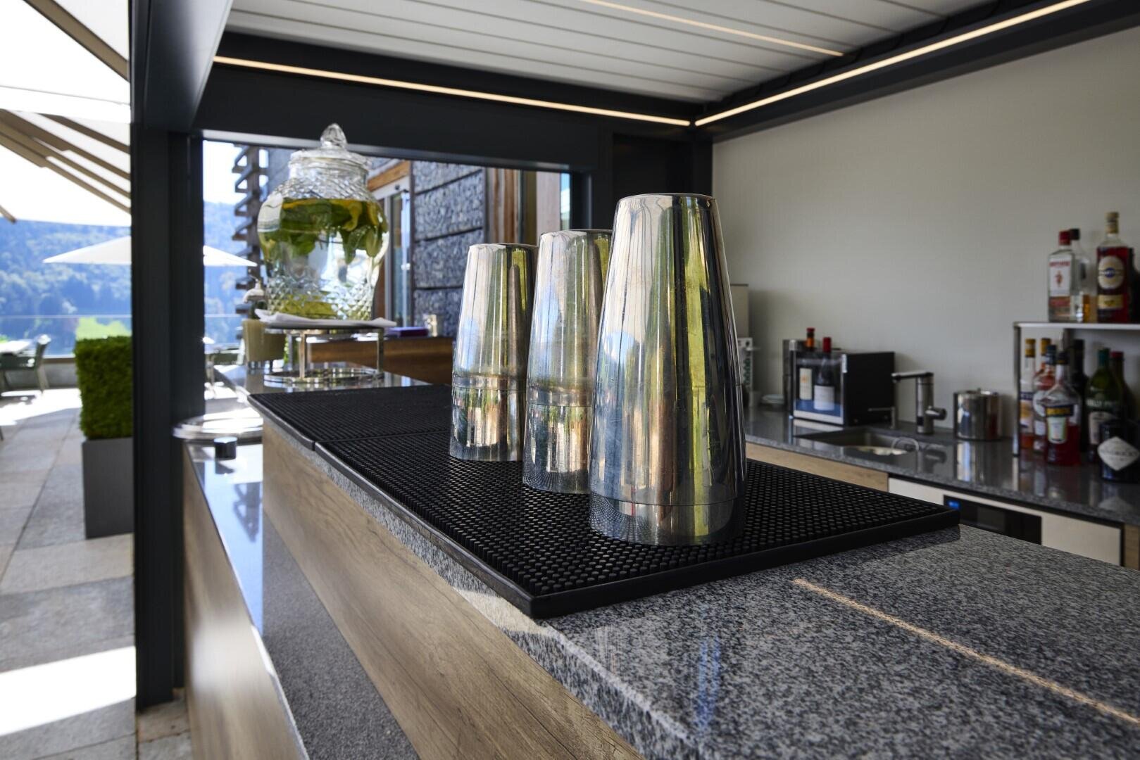 Eine Nahaufnahme von Metall-Cocktailshakern auf einer Granitarbeitsplatte an einer Bar im Freien, mit Flaschen und einem großen gläsernen Getränkespender im Hintergrund. Das Sonnenlicht beleuchtet die moderne Open-Air-Umgebung.