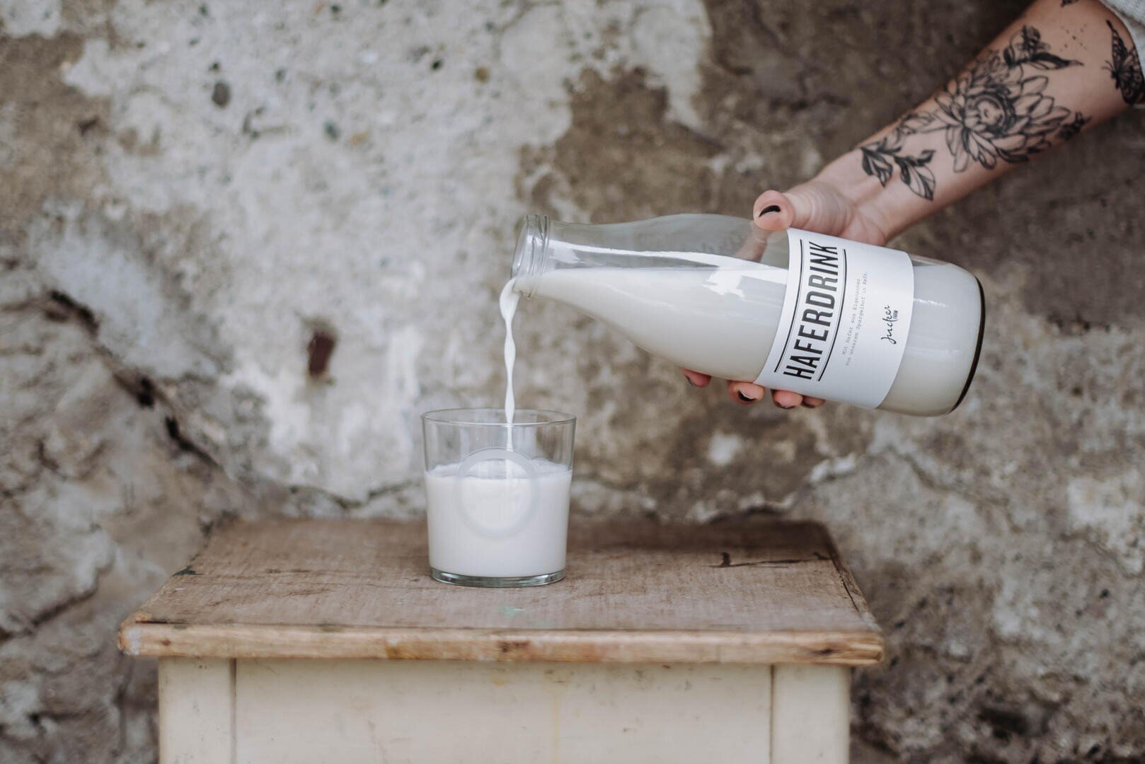 Ein tätowierter Arm gießt Hafermilch aus einer Glasflasche mit der Aufschrift Haferdrink in ein Glas auf einem kleinen Holztisch, mit einer strukturierten, verwitterten Wand im Hintergrund.