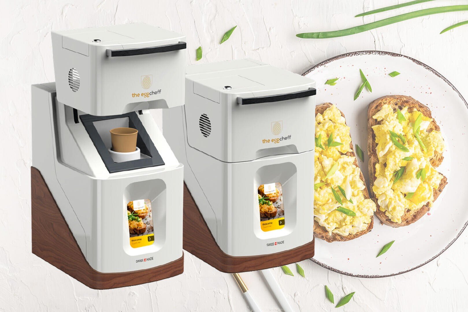 Zwei moderne weiße Küchengeräte mit digitalen Bildschirmen auf einer Arbeitsplatte, daneben ein Teller mit Rührei auf Toast, garniert mit grünen Kräutern. Neben dem Teller sind Essstäbchen platziert.