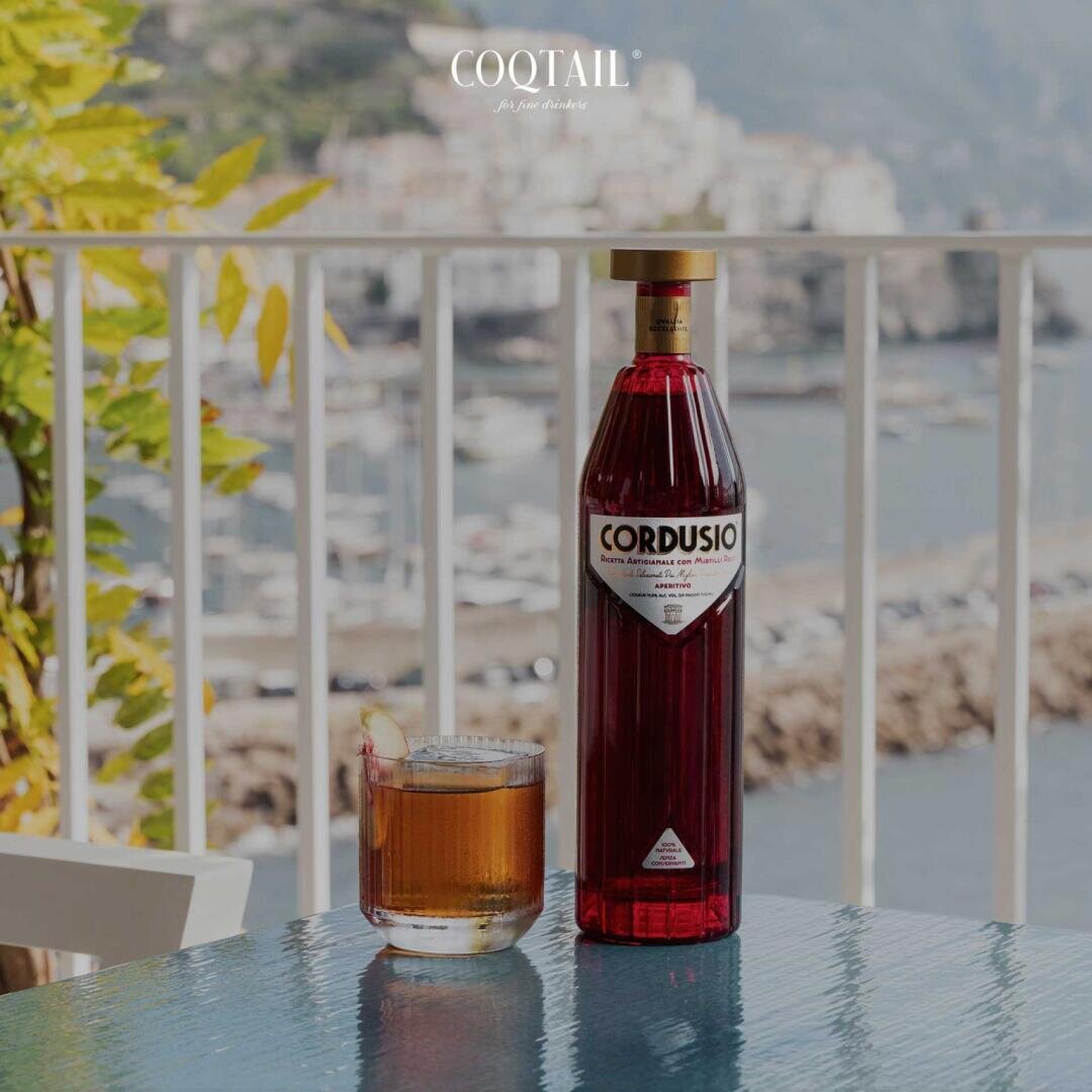 Eine Flasche Cordusio-Likör und ein Cocktailglas mit einem Zitronenzweig stehen auf einem blauen Tisch im Freien, mit Blick auf eine malerische Wasserlandschaft und Berge im Hintergrund. Am oberen Rand des Bildes steht COQTAIL geschrieben.