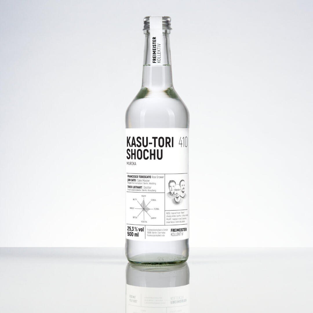 Une bouteille en verre transparent de KASU-TORI SHOCHU avec une étiquette blanche avec un texte et des illustrations en noir, qui se tient debout sur une surface réfléchissante sur un fond blanc simple.