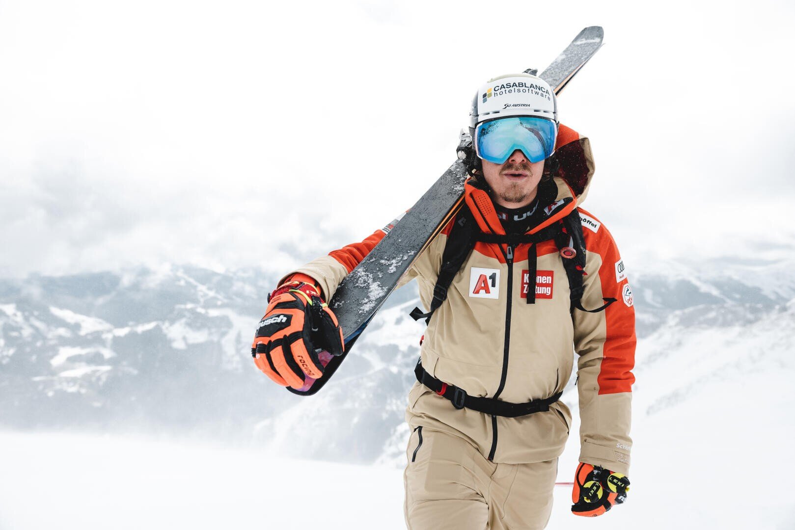 Ein Skifahrer in einem beige-roten Anzug mit Sponsorenlogos trägt Skier auf der Schulter, trägt einen Helm und eine blaue Skibrille und steht in einer verschneiten Berglandschaft.