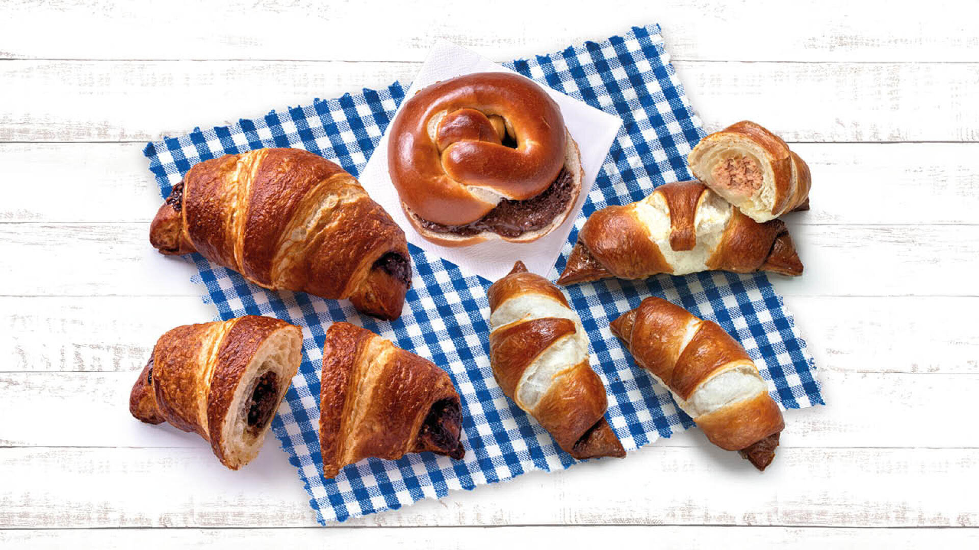 Eine Auswahl an Croissants und ein Brezelsandwich auf einem blau-weiß karierten Tuch auf einer weißen Holzfläche.