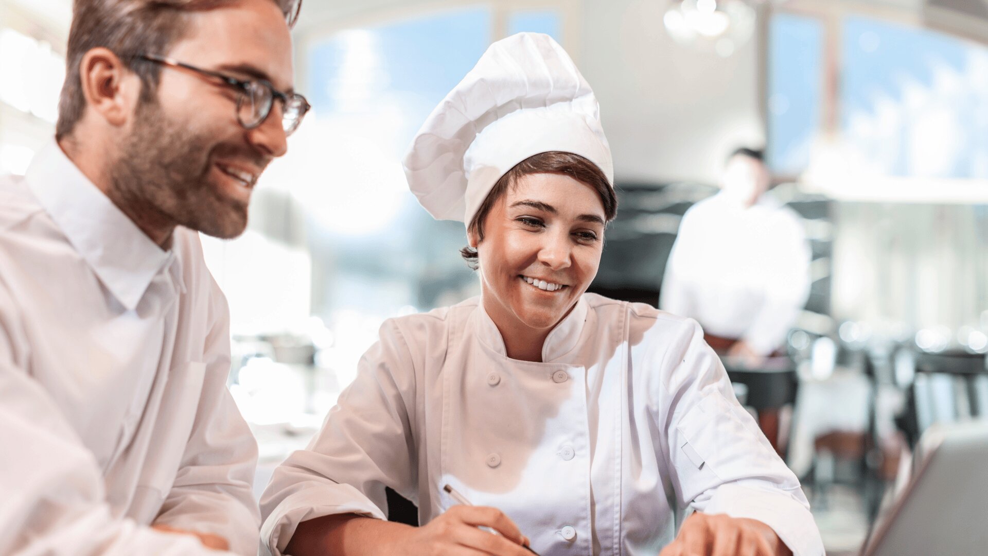 Zwei Köche in weißen Uniformen und Mützen sitzen zusammen an einem Tisch, lächeln und schauen auf etwas, das nicht zu sehen ist. Der Hintergrund zeigt ein helles, modernes Restaurant mit einer weiteren Person, die in der Ferne verschwimmt.