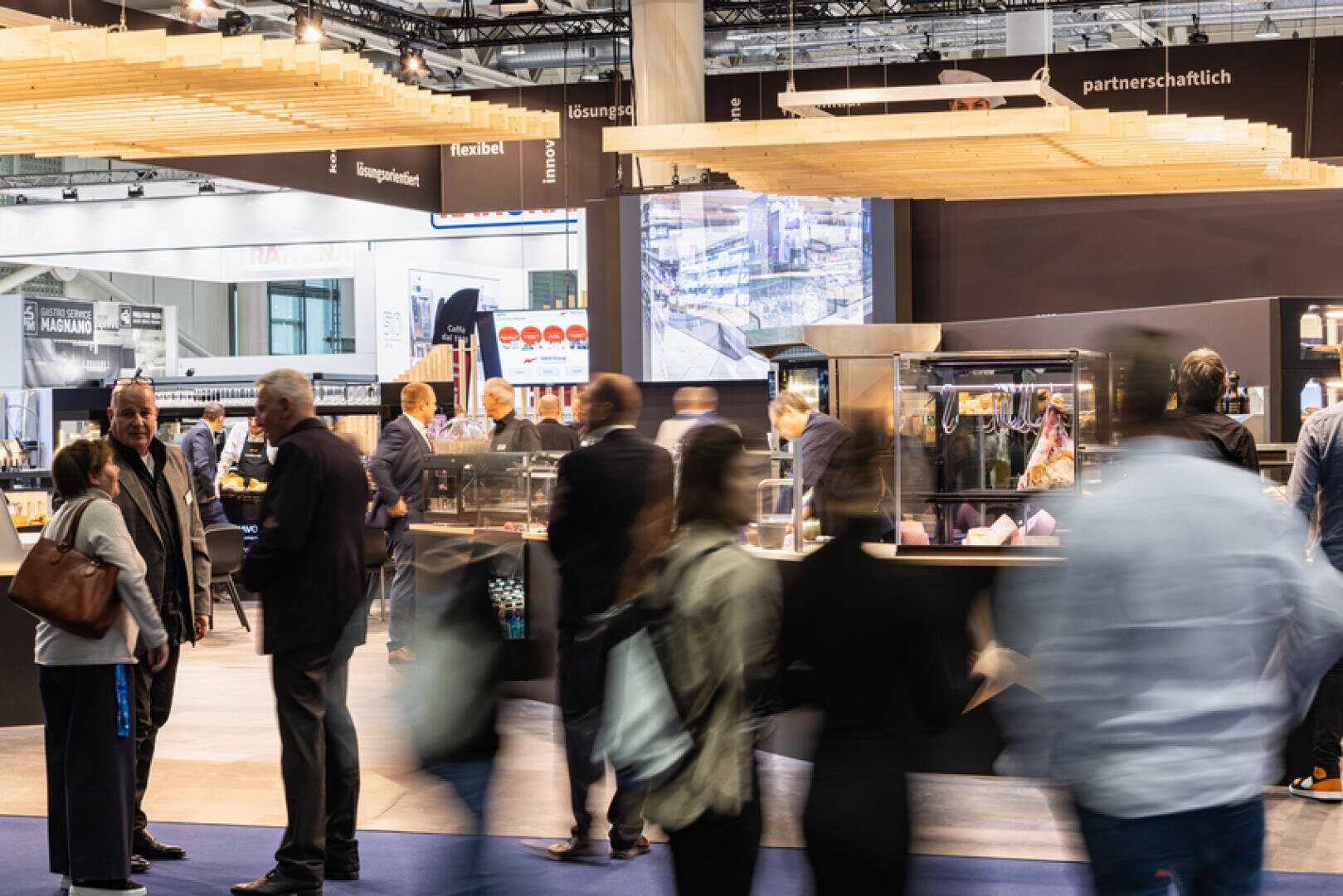 Menschen gehen um einen belebten Ausstellungsstand auf einer Messe herum, mit einer modernen Ausstellungsfläche, Informationsbildschirmen und hölzernen Dekorationsstrukturen über dem Kopf. Die Szene ist lebendig, mit unscharfen Bewegungen, die auf Bewegung hindeuten.