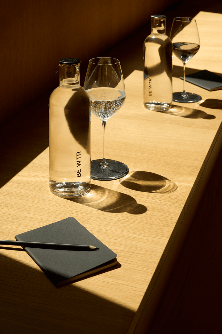 Deux bouteilles en verre remplies d'eau, deux verres à vin remplis d'eau et deux carnets de notes avec des crayons, disposés sur une surface en bois exposée au soleil et projetant de fortes ombres.