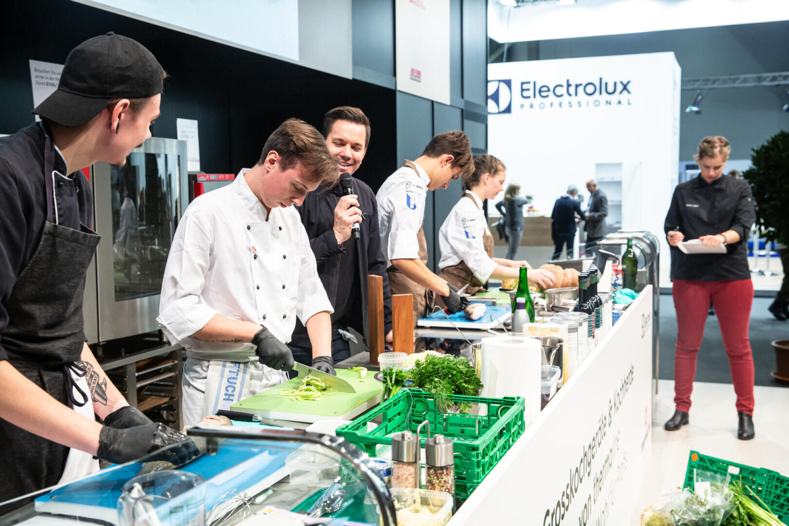 Eine Gruppe von Köchen bereitet während einer Kochvorführung an einer Theke Speisen zu, während ein Mann mit einem Mikrofon spricht und andere zusehen. Der Schauplatz scheint eine professionelle Küchenveranstaltung oder Ausstellung zu sein.