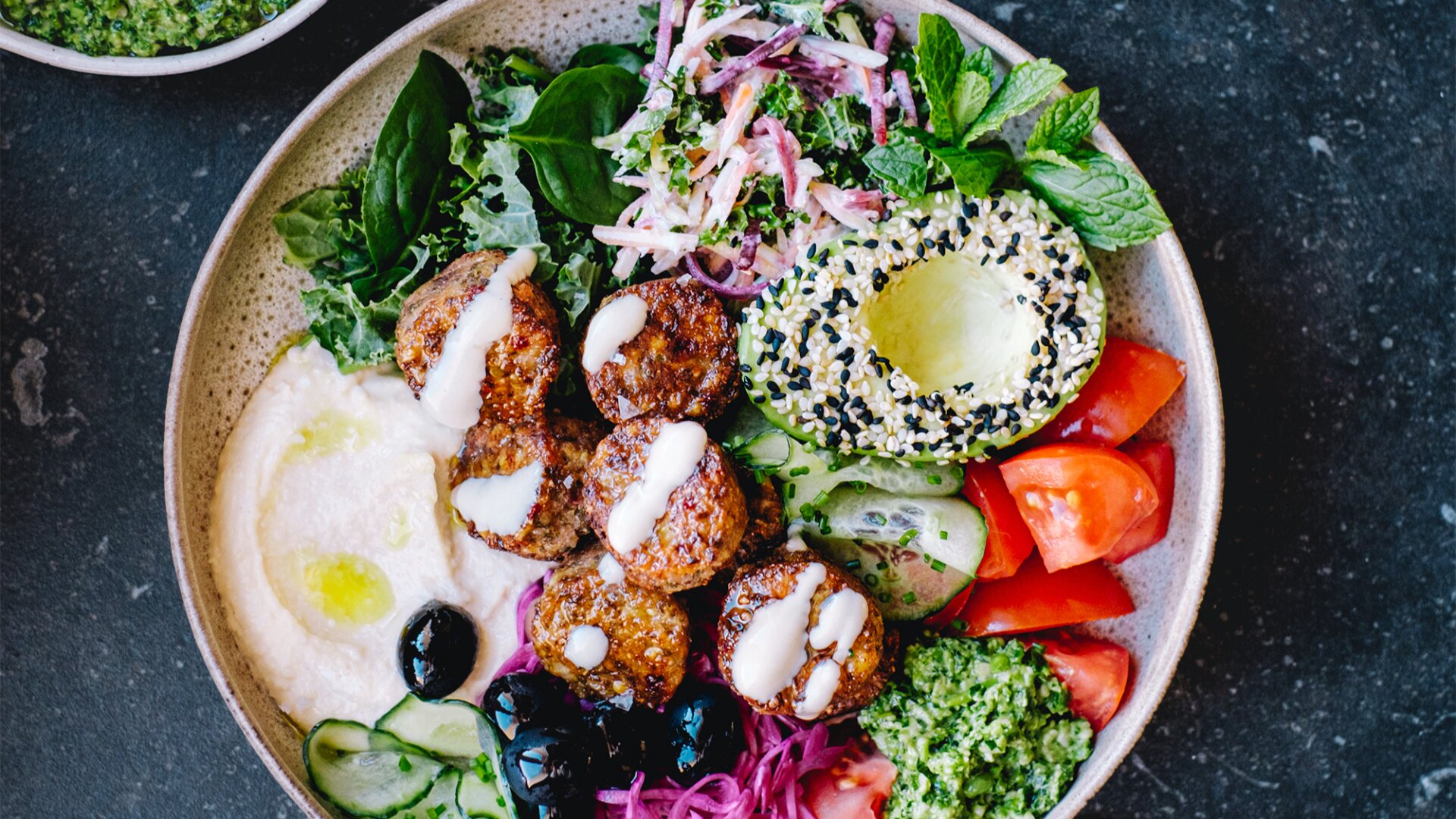 Eine Schale mit Falafel, Gurkenscheiben, Tomaten, Oliven, eingelegtem Gemüse, Hummus, Avocado mit Sesam und Kräutern, serviert mit grüner Soße und knusprigen Bällchen aus Mungbohnen auf einem dunklen Steinuntergrund.
