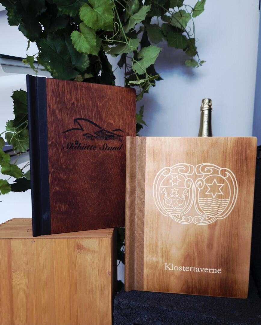 Zwei holzverkleidete Speisekarten stehen aufrecht neben einer Flasche Champagner und grünen Pflanzen. Auf einer Speisekarte steht Schöne Strand mit einer Illustration, auf der anderen Klostertaverne mit einem verschnörkelten Kreis.