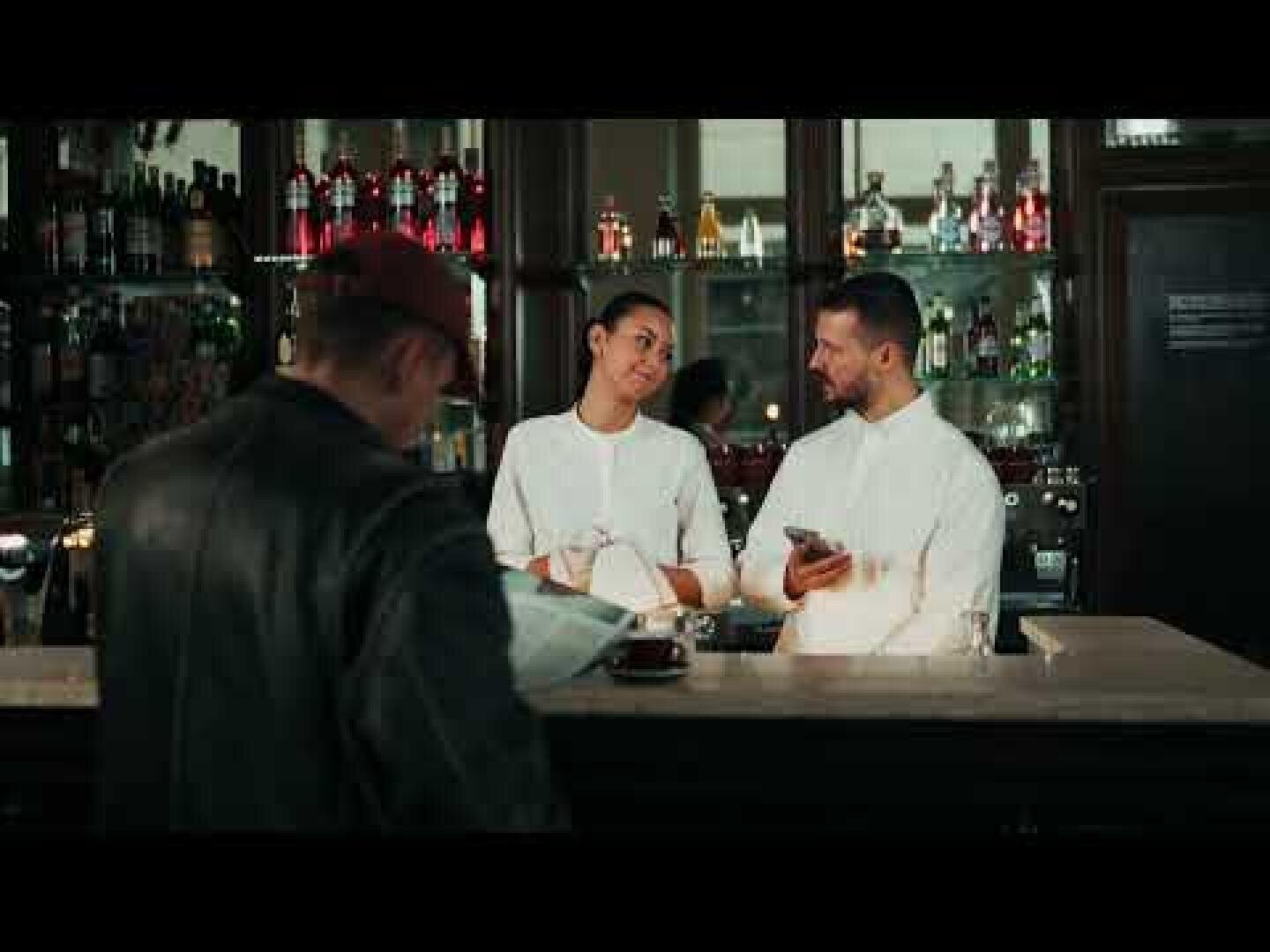 Zwei Barkeeper in weißen Uniformen stehen hinter einer Bar, unterhalten sich und lächeln. Hinter ihnen sind Flaschen in Regalen ausgestellt. Im Vordergrund liest eine Person mit Hut an der Bar eine Zeitung.