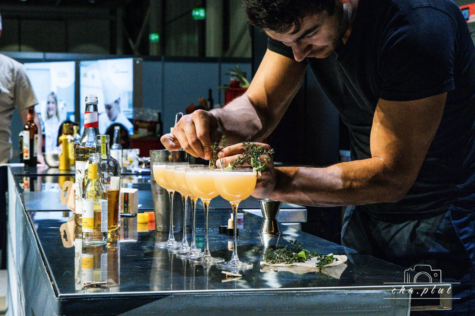 Un barman décore soigneusement une série de cocktails jaune-orange sur un comptoir métallique brillant, entouré de bouteilles, d'ingrédients et d'outils à cocktails lors d'un événement ou d'un concours animé.