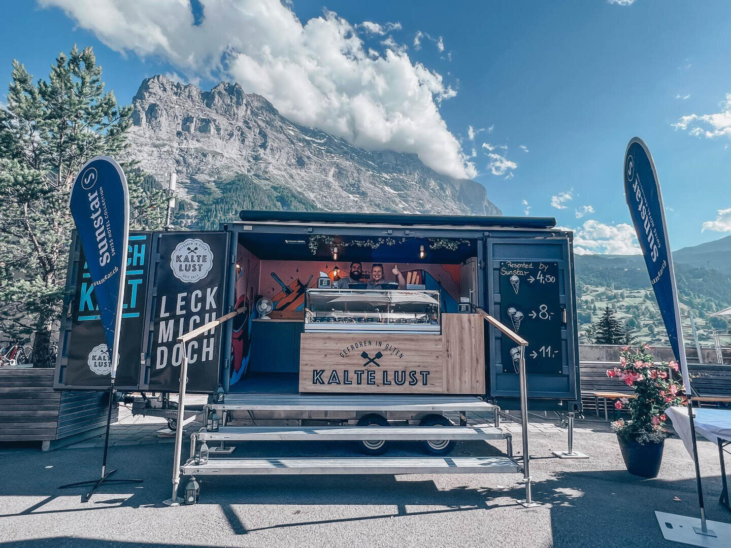 Ein Eiswagen mit dem Namen Kalte Lust und Personal im Inneren steht vor einer malerischen Bergkulisse. Auf beiden Seiten sind blaue Banner angebracht, und auf dem Wagen ist eine Speisekarte abgebildet. Der Himmel ist teilweise bewölkt.