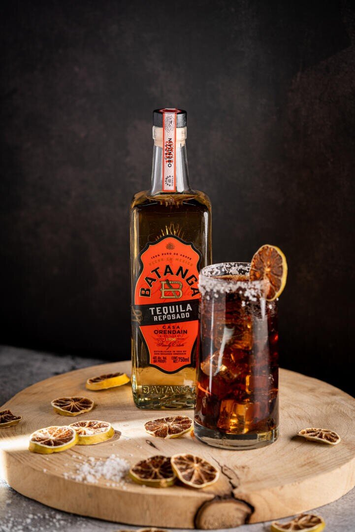 Eine Flasche Batanga Tequila Reposado steht neben einem hohen Glas eines dunklen Cocktails mit Eis und einer Garnitur aus getrockneten Zitrusfrüchten, die auf einem Holzbrett mit verstreuten getrockneten Zitrusscheiben vor einem dunklen Hintergrund stehen.