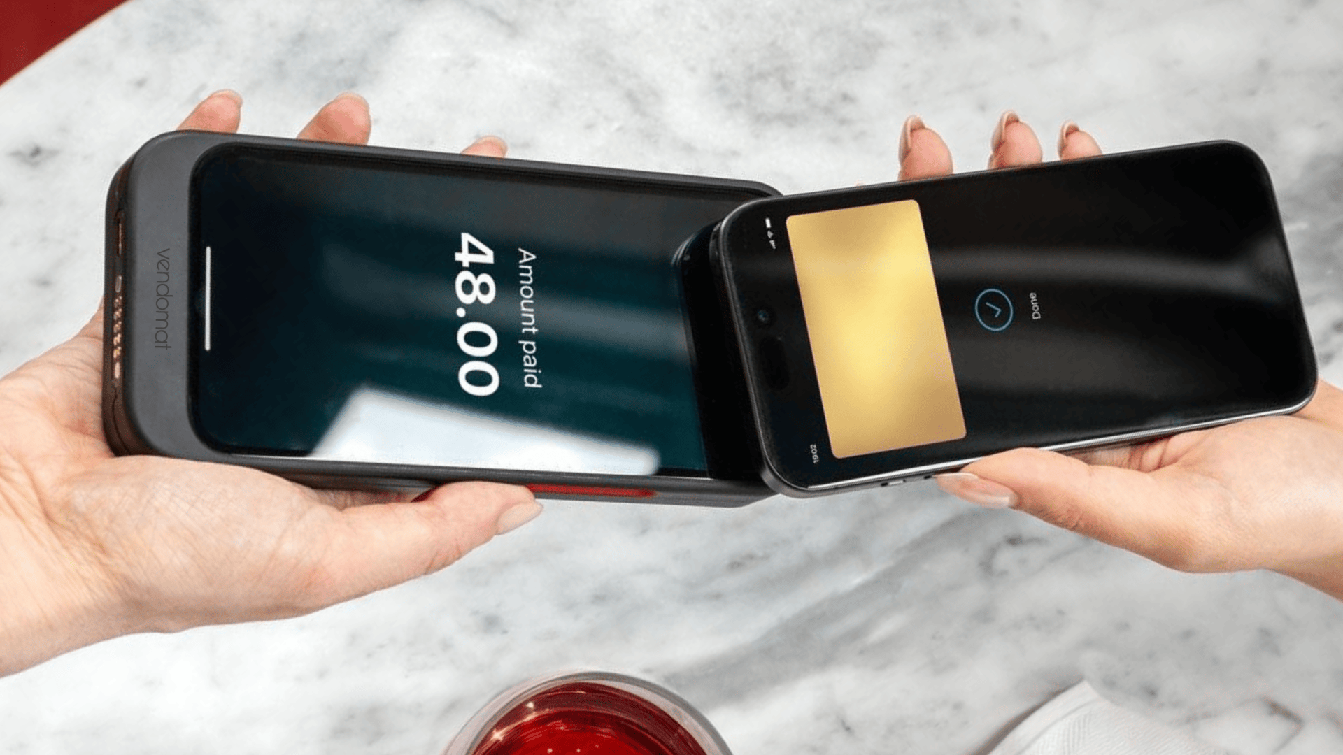 Zwei Hände halten ein Zahlungsterminal und ein Smartphone und führen eine kontaktlose Zahlung von 48,00 durch. Die Geräte berühren sich über einer Marmoroberfläche, und ein rotes Getränk ist teilweise darunter zu sehen.