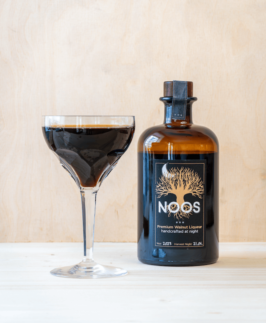 Ein mit dunklem Walnusslikör gefülltes Glas steht neben einer braunen Flasche mit der Aufschrift "NOOS Premium Walnut Liqueur handcrafted at night" auf einer hellen Holzoberfläche mit einem schlichten hellen Hintergrund.