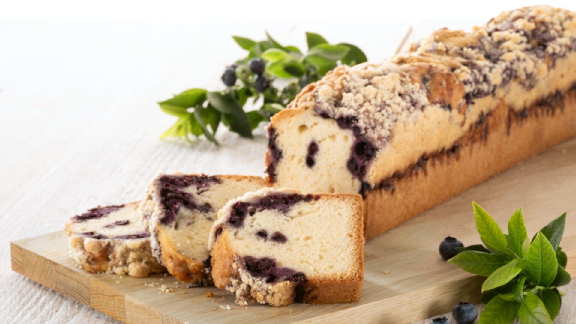 Ein Laib Blaubeer-Streuselkuchen auf einem Holzbrett, mit zwei geschnittenen und ausgestellten Scheiben. Frische Blaubeeren und grüne Blätter werden zur Dekoration herumgelegt.