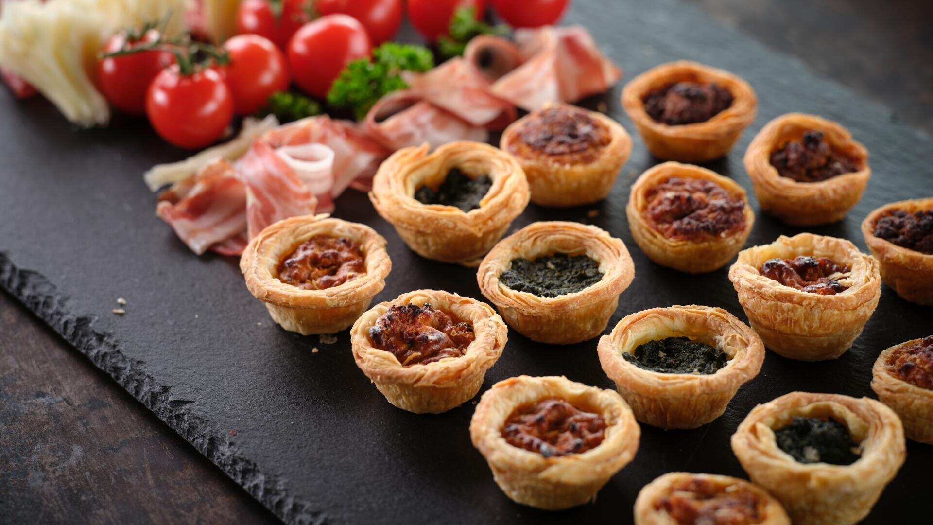 Eine Nahaufnahme von verschiedenen Mini-Quiches, die auf einer dunklen Schieferplatte angeordnet sind, mit Prosciuttoscheiben, Käse, Kirschtomaten und Petersilie im Hintergrund.