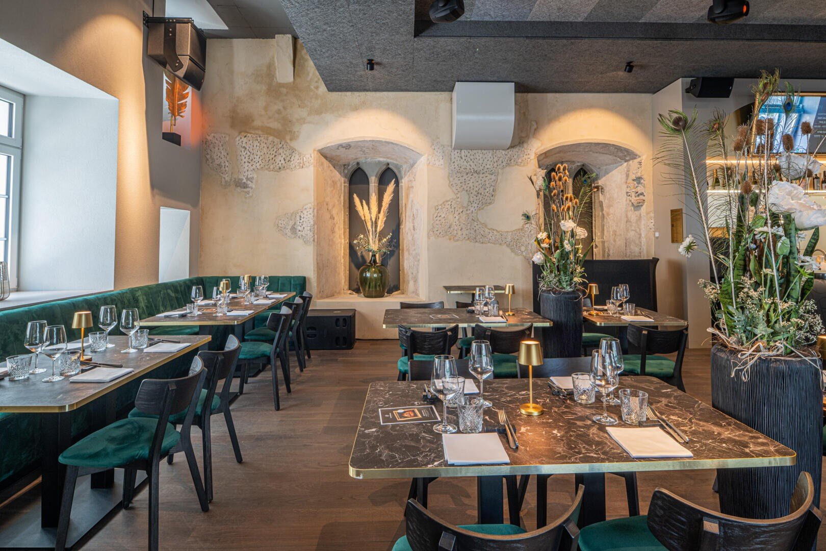 Ein stilvolles Restaurantinterieur mit grünen Samtstühlen, schwarzen Marmortischen mit Glasgeschirr und dekorativen Pflanzen. Freiliegende strukturierte Wände und sanfte Beleuchtung schaffen eine moderne, einladende Atmosphäre.