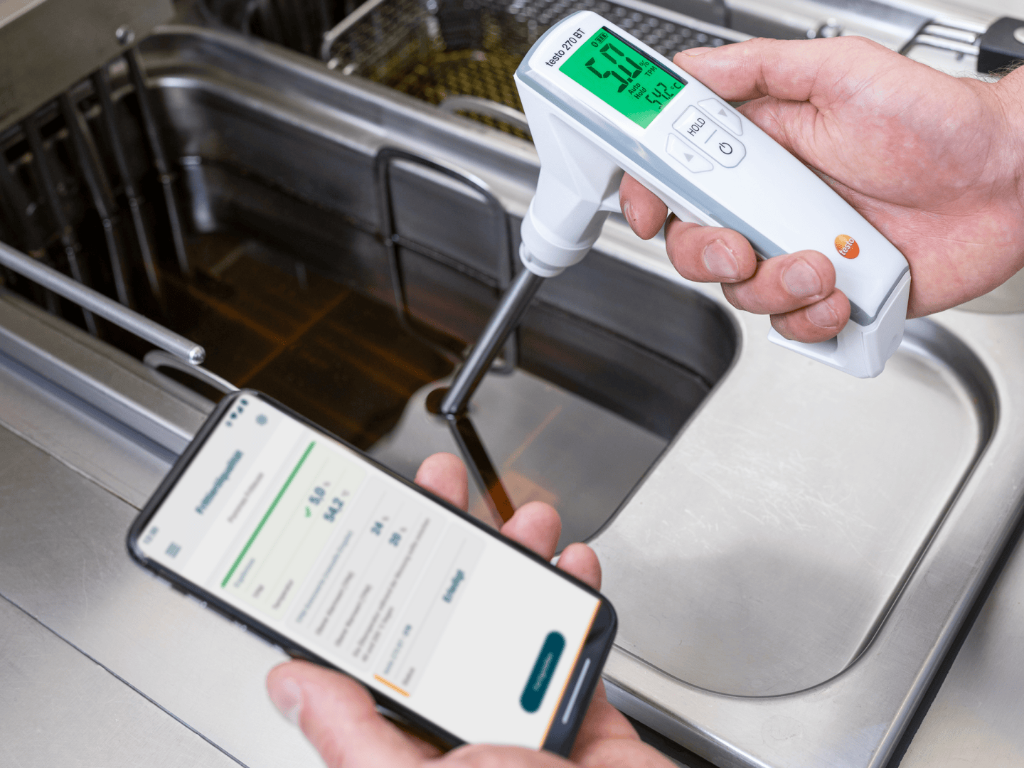 Eine Person verwendet ein tragbares Digitalthermometer, um die Temperatur des Öls in einer Fritteuse zu messen, während sie ein Smartphone in der Hand hält, das die Messdaten anzeigt.